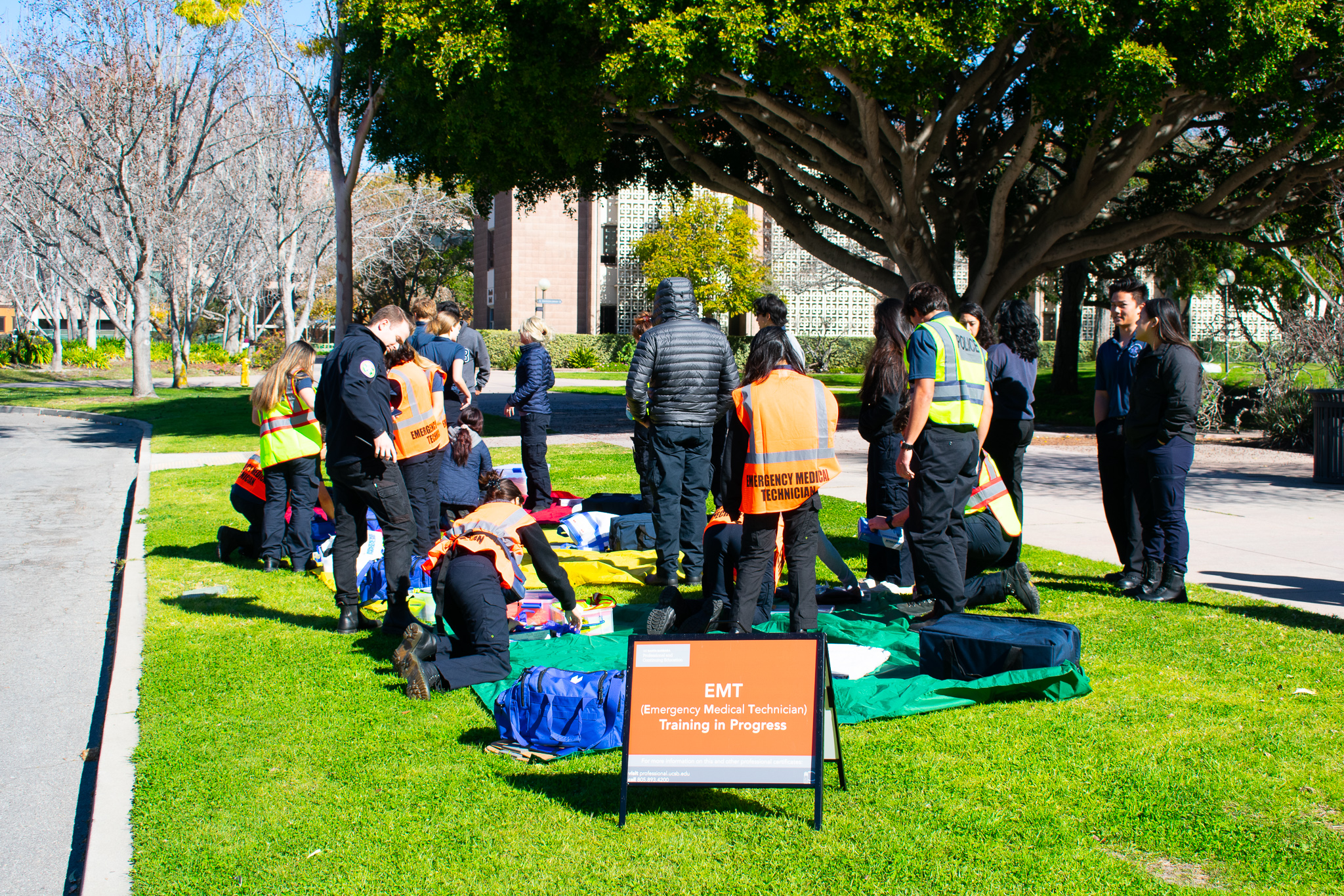 EMT Students kneeling down to work on a patient, in the foreground is an A-frame saying "EMT Training in Progress"